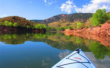 First-person view from the bow of a kayak on a calm reservoir reflecting green trees and red rock shorelines under a blue sky.